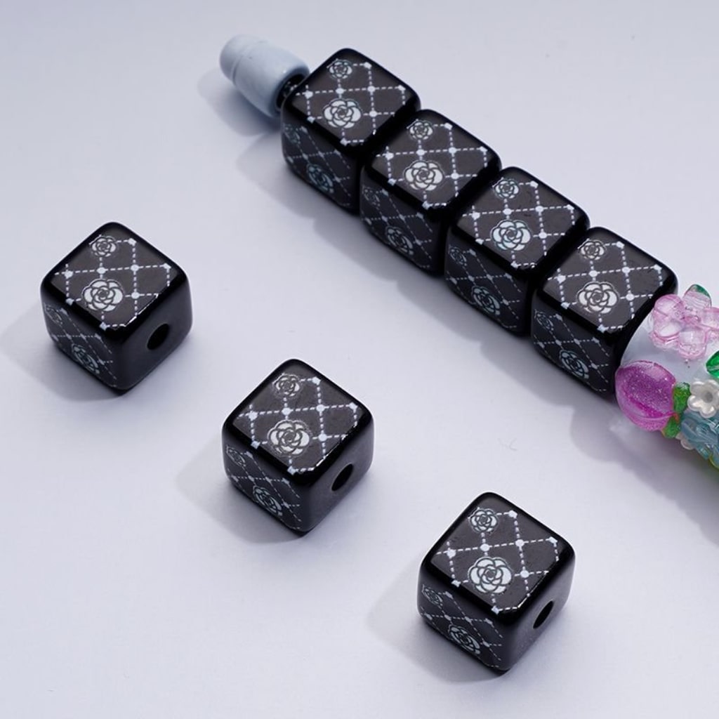 A series of black square cube acrylic beads with white rose flowers designs, displayed alongside other decorative items.