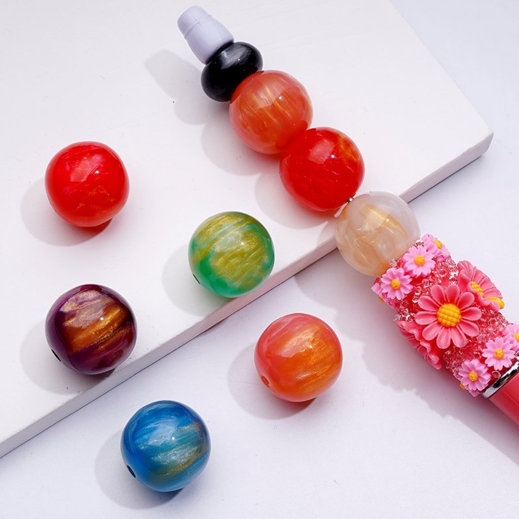 A selection of colorful round acrylic beads in various colors displayed alongside a pen with a bead attached to its cap.


