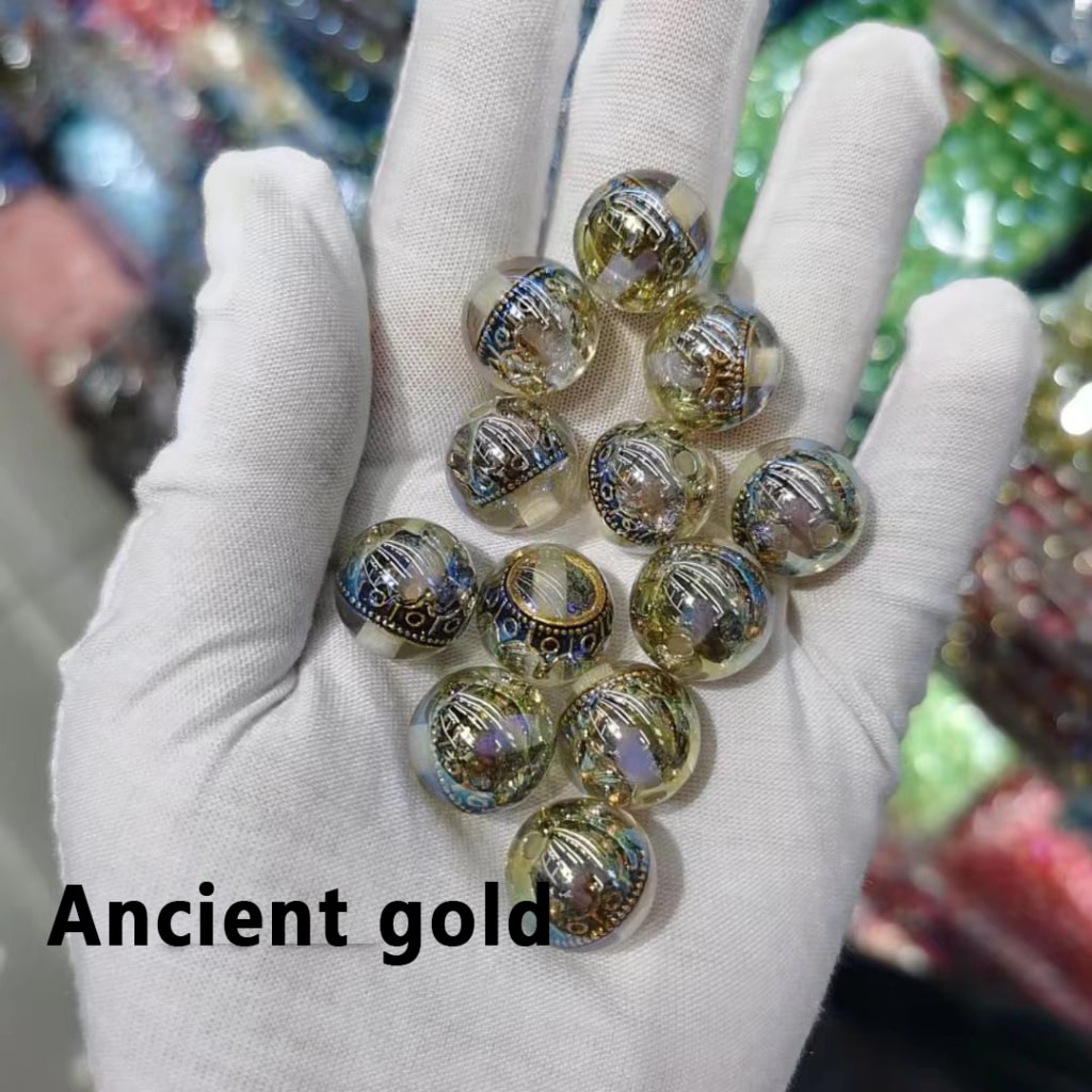 Hand wearing a white glove holding decorative beads labeled 'Ancient gold' against a blurred background.

