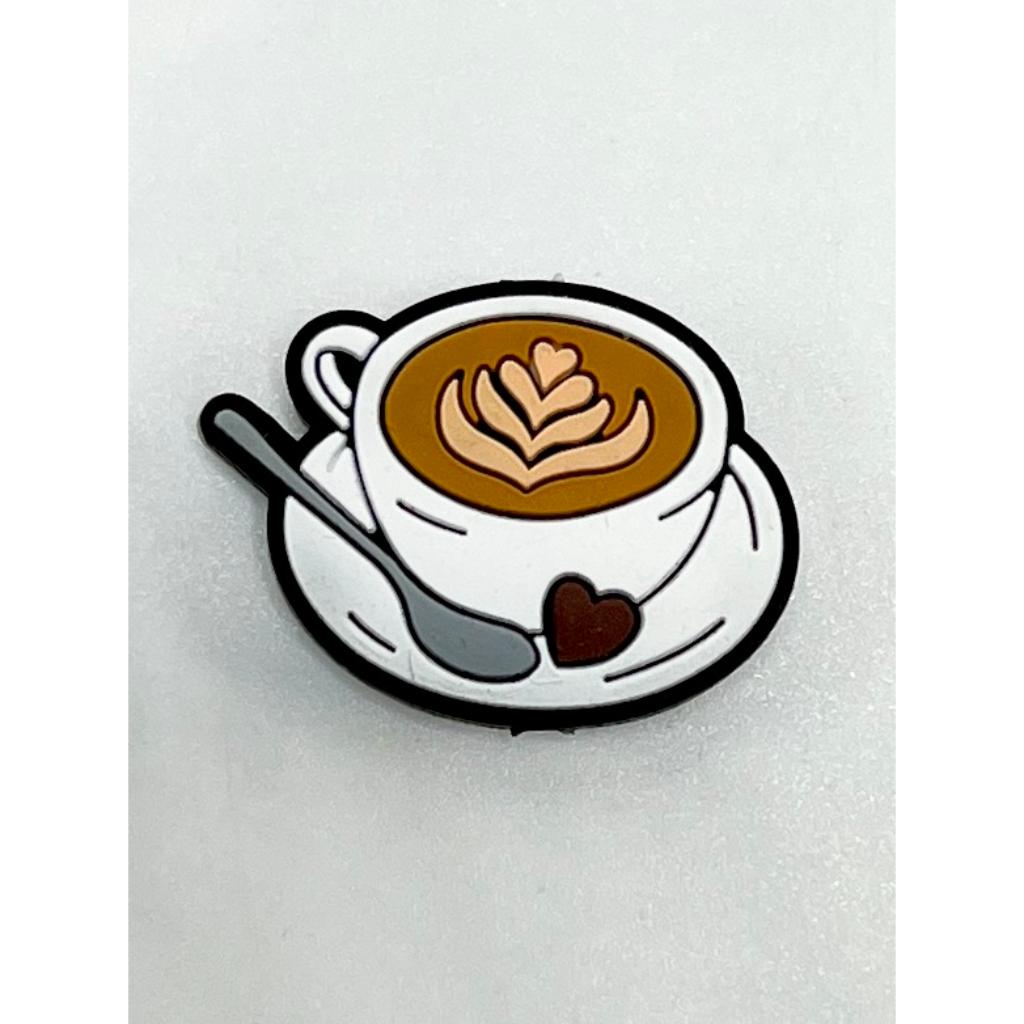 A silicone bead in the shape of a coffee cup with a latte art design on it, accompanied by a spoon, predominantly white with brown and black details.

