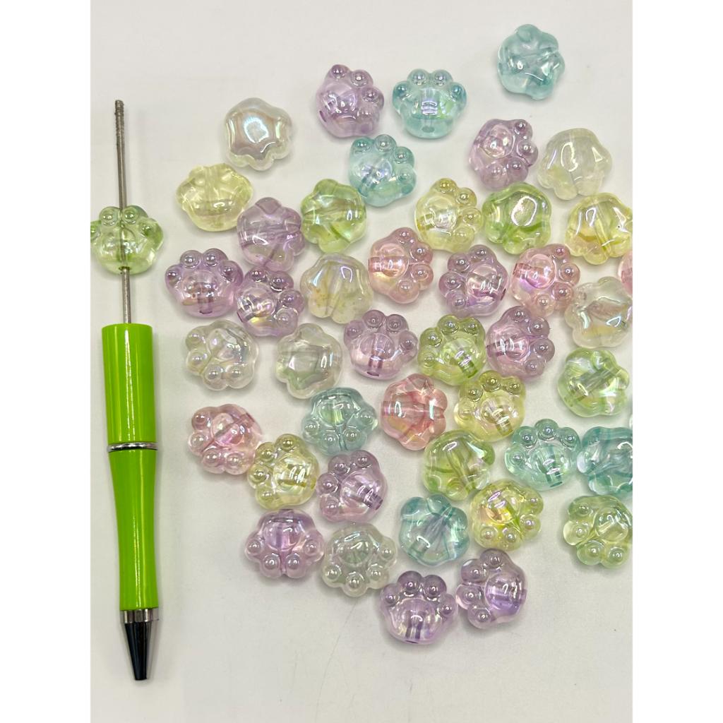 A collection of translucent acrylic beads in pink, purple, and clear colors, alongside a green bead threading tool with a needle.


