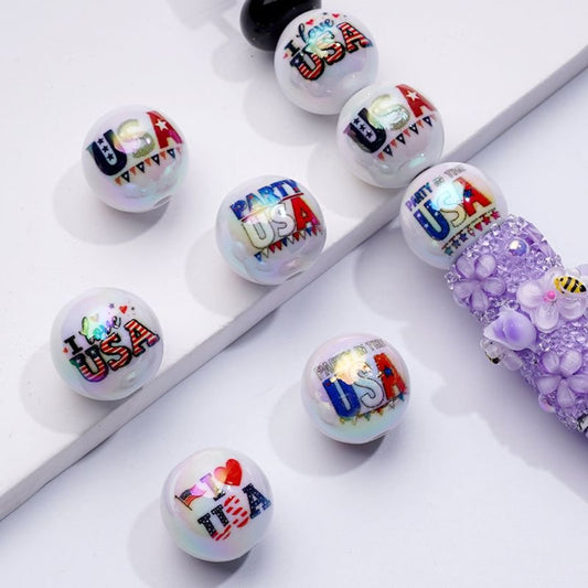 A collection of white round acrylic beads with embedded USA national flags and other patriotic designs, displayed on a surface.

