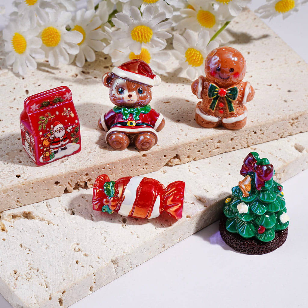 Five miniature Christmas-themed decorative figurines and a small red tin arranged on layered beige stone slabs with white daisies in the background. The set includes a Santa-themed tin, a teddy bear in a Santa hat, a gingerbread man, a wrapped candy, and a small Christmas tree.

