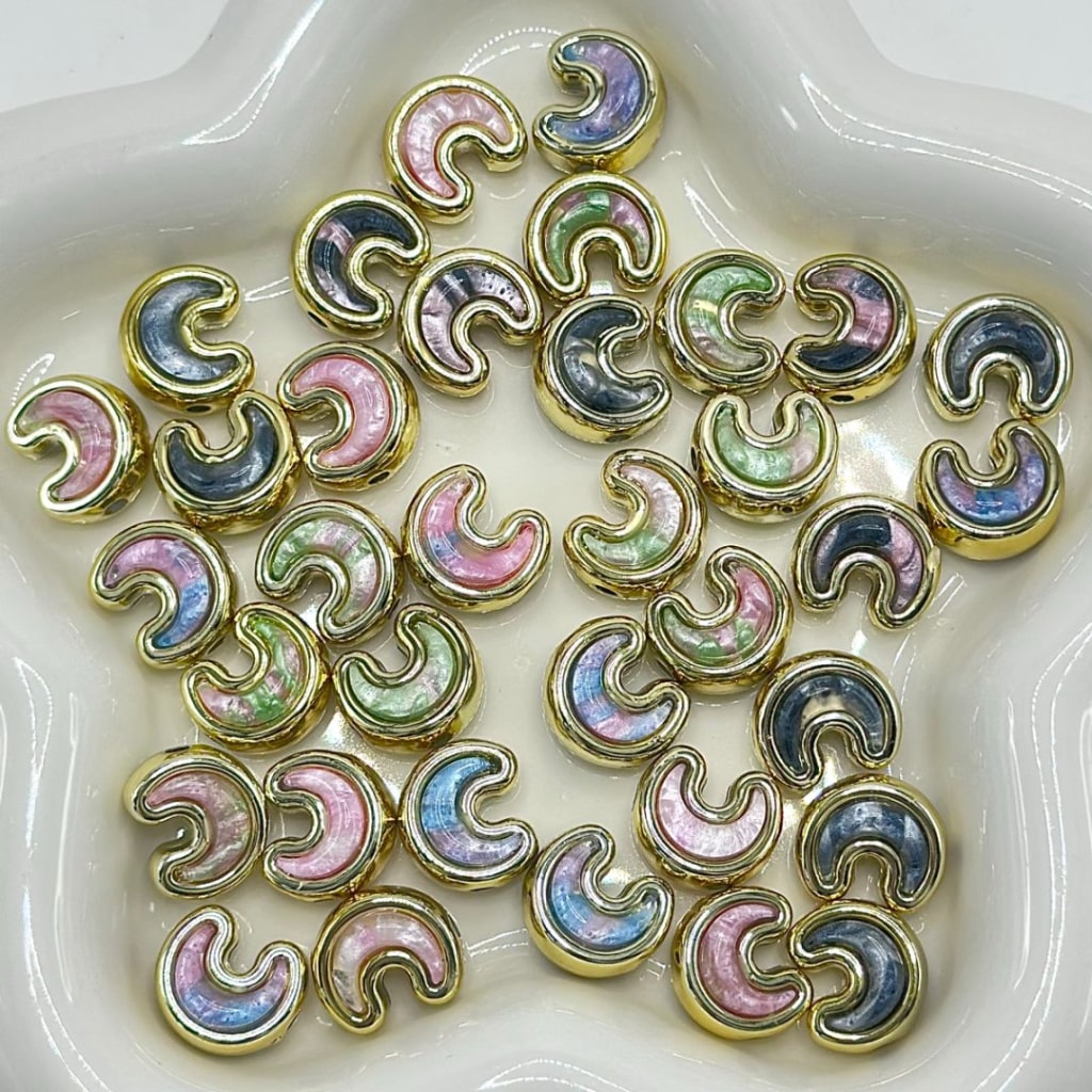 A group of small crescent-shaped jewelry charms with iridescent pastel inlays and gold-tone edges, arranged in a white star-shaped dish.

