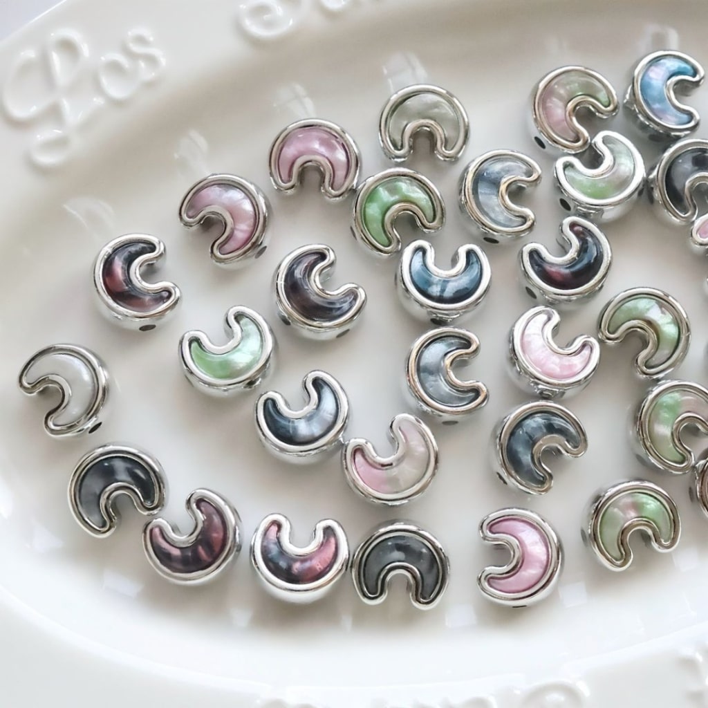 A collection of silver crescent-shaped beads with iridescent mother-of-pearl inlays in pastel colors, arranged on a white ceramic dish.

