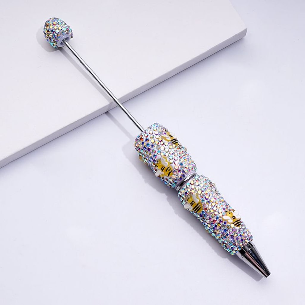A multicolored beadable clay pen with yellow bee and AB rhinestone detailing, placed on a white surface.

