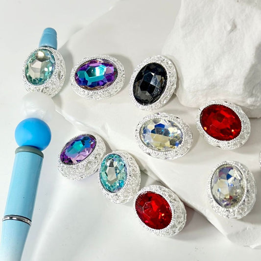 Assorted large shiny oval rhinestones with silver alloy hollow beads, displayed on a white surface next to a blue tool.

