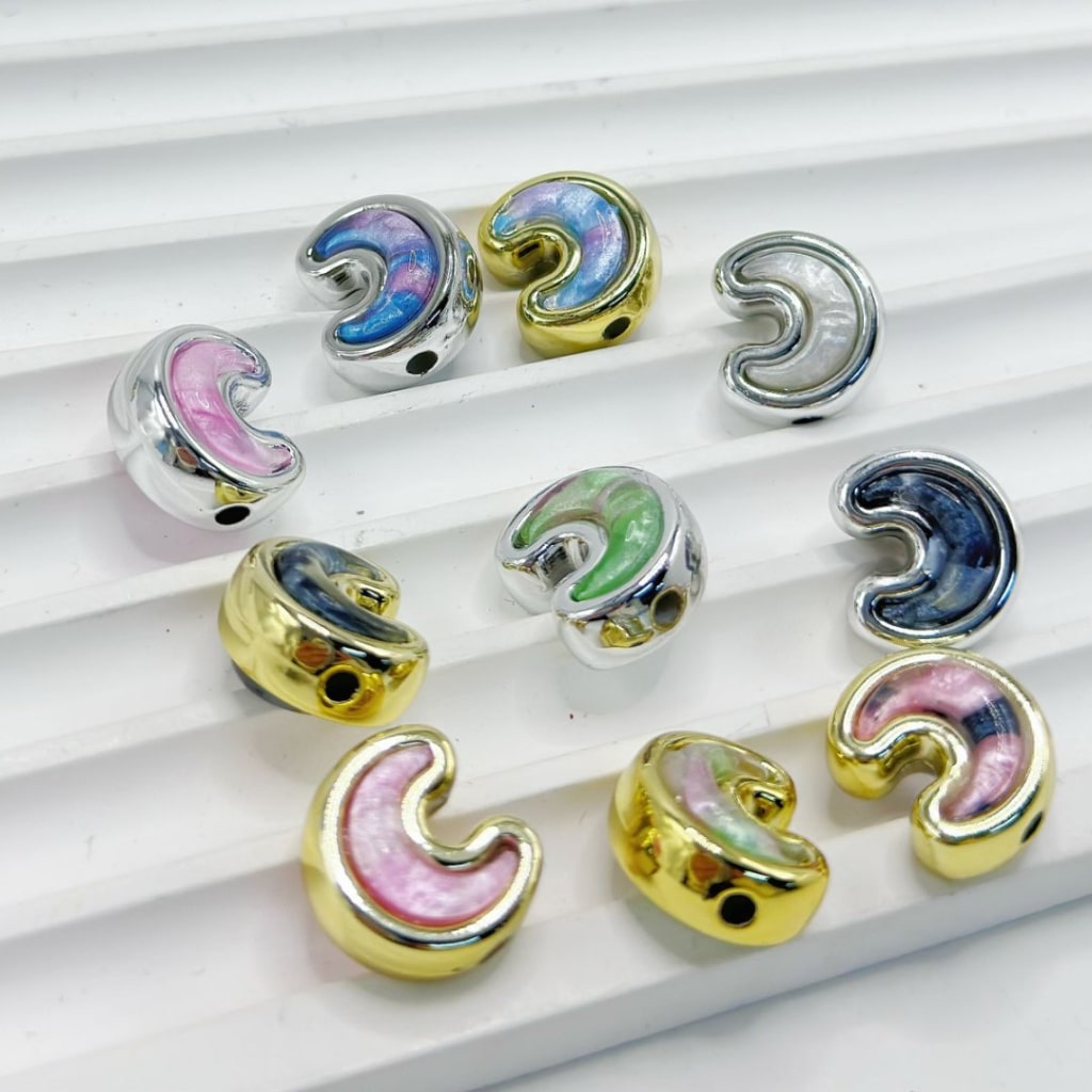 Nine metallic crescent-shaped beads with iridescent pastel fillings in pink, blue, green, and silver, arranged on a white ribbed surface.

